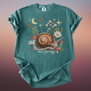 May include: Teal-colored t-shirt featuring a whimsical illustration. A large snail with a brown shell is surrounded by flowers, mushrooms, and butterflies. The text "Work Your Magic" is printed below the snail. The shirt has a crew neck and short sleeves.