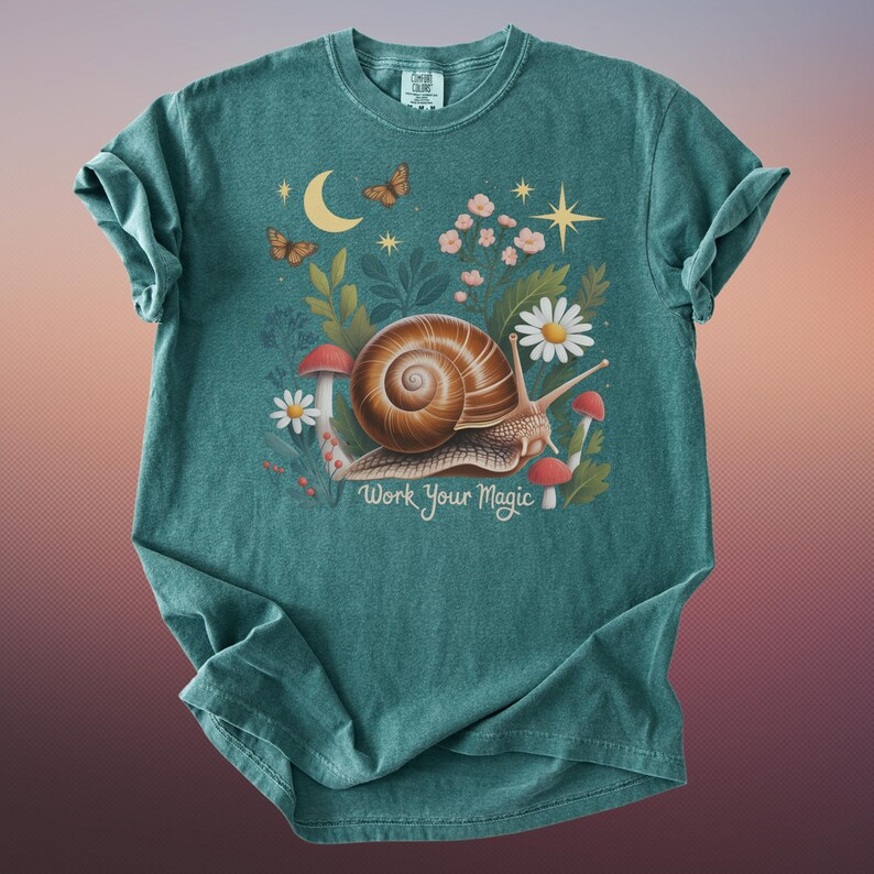 May include: Teal-colored t-shirt featuring a whimsical illustration. A large snail with a brown shell is surrounded by flowers, mushrooms, and butterflies. The text "Work Your Magic" is printed below the snail. The shirt has a crew neck and short sleeves.