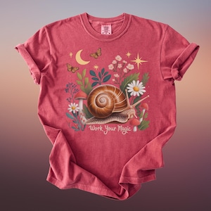 May include: A red t-shirt featuring a whimsical illustration of a snail with a spiral shell, surrounded by flowers, mushrooms, butterflies, and a crescent moon. The text "Work Your Magic" is below the snail.