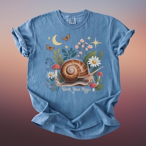 May include: A light blue t-shirt featuring a whimsical illustration of a snail with a brown shell, surrounded by flowers, mushrooms, butterflies, and a crescent moon. The text "Work Your Magic" is below the snail.