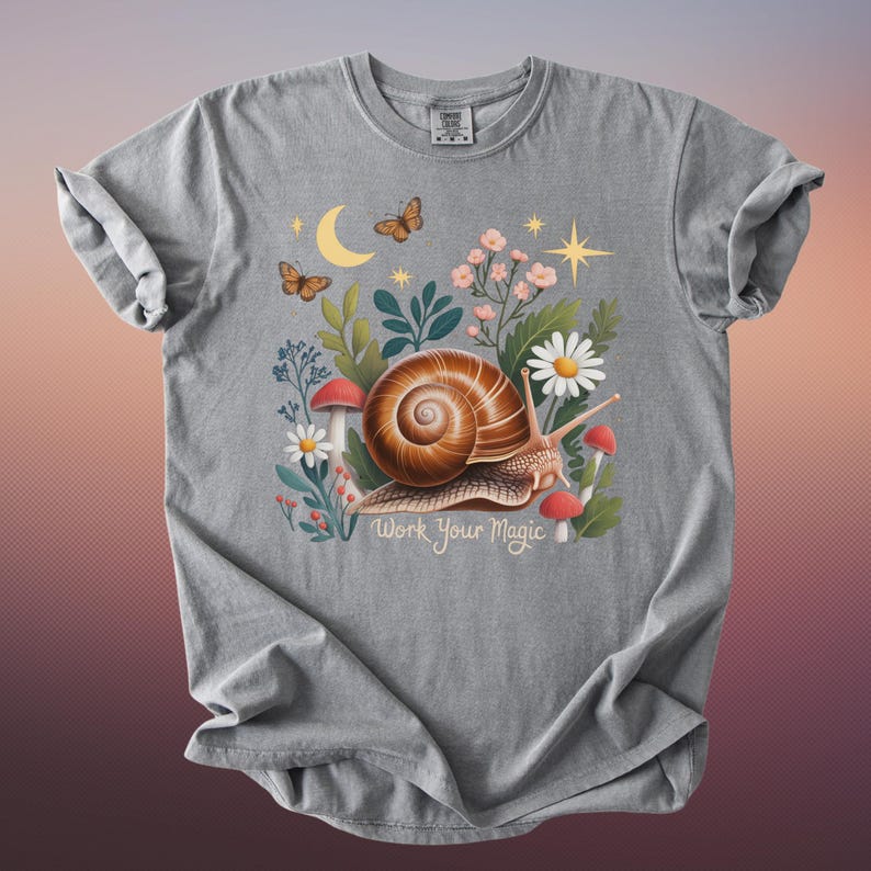 May include: Gray t-shirt featuring a whimsical illustration of a snail with a spiral shell, surrounded by flowers, mushrooms, butterflies, and a crescent moon. The text "Work Your Magic" is below the snail.