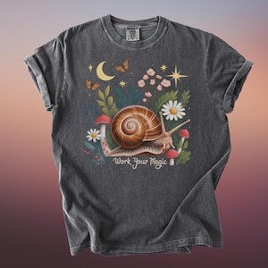 May include: Dark gray t-shirt featuring a whimsical illustration of a snail with a brown shell, surrounded by mushrooms, flowers, and butterflies. The text "Work Your Magic" is below the snail. The shirt has rolled-up sleeves.