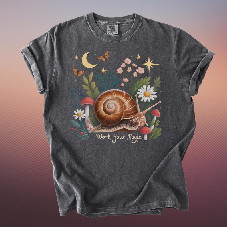 May include: Dark gray t-shirt featuring a whimsical illustration of a snail with a brown shell, surrounded by mushrooms, flowers, and butterflies. The text "Work Your Magic" is below the snail. The shirt has rolled-up sleeves.