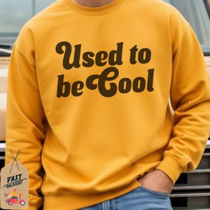May include: Mustard yellow sweatshirt with the text "Used to be Cool" in a brown, retro font. The sweatshirt has a crew neck and long sleeves. The person is wearing blue jeans.