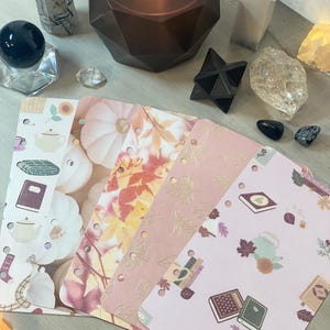 May include: A collection of decorative crystals, a black star, and a lit candle in a geometric holder. Several planner inserts with fall-themed patterns, including pumpkins, leaves, and books, are fanned out on a light-colored surface.