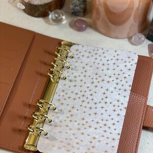 May include: A brown leather planner with gold-colored rings and a white insert with a gold star pattern. The planner is open, revealing the insert. A lit candle and decorative crystals are in the background.