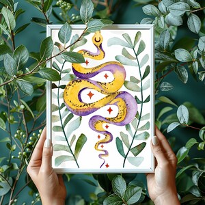 Op de afbeelding: Een ingelijste kunstdruk met een aquarelillustratie van een kronkelende slang in tinten geel en paars. De slang is omringd door groene bladeren en versierd met kleine rode sterren en een maansikkel-symbool boven de kop van de slang.