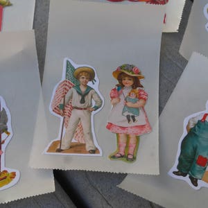 May include: Paper bags decorated with vintage stickers. The stickers depict children in patriotic attire, including sailor suits and overalls, holding flags and dolls, and a firework display with the text "4th".