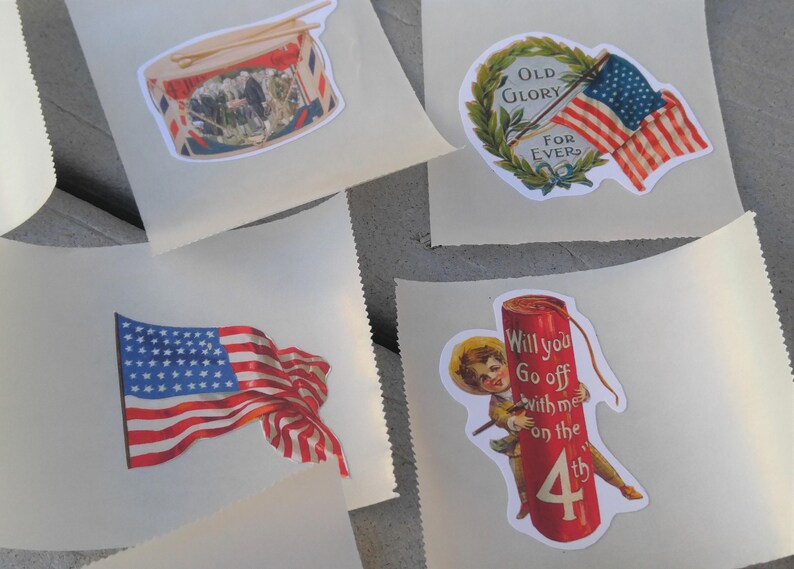 May include: A selection of vintage-style stickers with patriotic designs. The stickers depict an American flag, a drum, and a firecracker with the text "Will you go off with me on the 4th". They are presented on a light-coloured background.