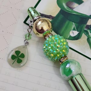 May include: A green and gold beaded pen with a four-leaf clover charm. The pen features a green tip, a gold sphere, a green beaded sphere, and a clear teardrop charm with a green clover. The pen is a St. Patrick's Day themed item.
