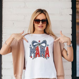 May include: White t-shirt with a patriotic "MAMA" design. The design features a blue bow with stars, red and blue accents, and the word "MAMA" in red letters. The shirt is a casual style.