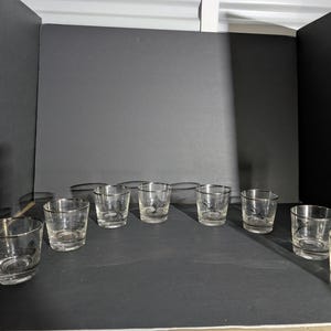 May include: A collection of clear glass tumblers with silver rims. Some tumblers have etched designs. The glasses are arranged on a dark surface against a dark background. The glasses are empty.