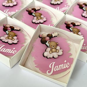 Può includere: Diversi cerchi in acrilico trasparente con un design a nuvola rosa, un orsacchiotto e il nome "Jamie" in corsivo bianco sono presentati in scatole bianche. L'orsacchiotto tiene in mano dei palloncini.