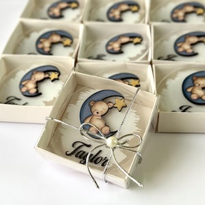 May include: Baby shower favours presented in small boxes. Each favour features a teddy bear on a crescent moon with a star. The boxes are tied with silver ribbon and a pearl. The name "Taylor" is printed on the favours.