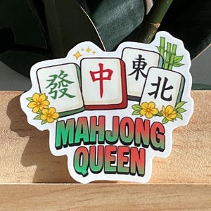 May include: A sticker with the text "MAHJONG QUEEN" in green and white, with three Mahjong tiles. The tiles have Chinese characters and are surrounded by yellow flowers. The sticker is on a wooden surface.