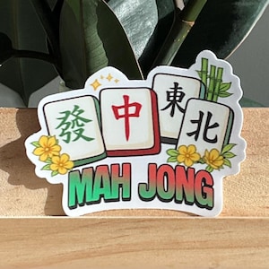 May include: A colorful sticker featuring Mah Jong tiles with Chinese characters and the words "MAH JONG" in green. The sticker is decorated with yellow flowers and is placed on a wooden surface, with a leafy green plant in the background.