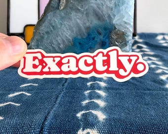 Op de afbeelding: Een gestanste sticker met het woord Exactly. in een vet, rood retro-lettertype met een witte omlijning. De sticker is geplaatst voor een blauwe agaatplak en een glas-in-loodpaneel met een bloemmotief op een blauwe stof met patroon.