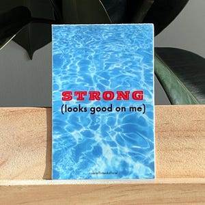 May include: A rectangular sticker with a blue water pattern and the word "STRONG" in red, with the phrase "(looks good on me)" below it. The sticker is displayed on a wooden surface with a green plant in the background.