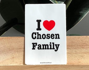 Op de afbeelding: Een witte rechthoekige sticker met de tekst "I ♥ Chosen Family" in zwart en een rood hartsymbool. De sticker is geplaatst op een houten oppervlak, met een plant op de achtergrond.