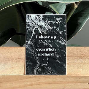 May include: A rectangular sticker with a black and white marble design. The sticker has the text "I show up even when it's hard" in a white, sans-serif font. The sticker is displayed on a wooden surface, with green leaves in the background.
