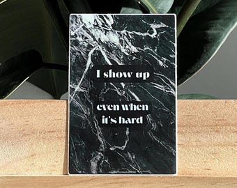 Op de afbeelding: Een rechthoekige sticker met een zwart-wit marmerontwerp. De sticker heeft de tekst "I show up even when it's hard" in het wit. De sticker wordt weergegeven op een houten oppervlak, met groene bladeren op de achtergrond.