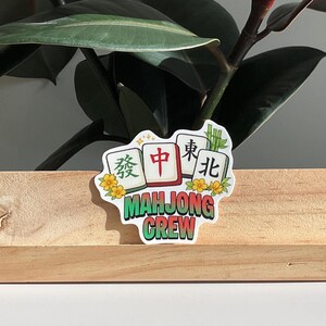 May include: A sticker with the text "MAHJONG CREW" in green, with Chinese characters on white tiles. The sticker has a floral design and is set against a wooden surface and green plant.