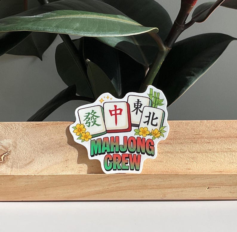 May include: A sticker with the text "MAHJONG CREW" in green, with Chinese characters on white tiles. The sticker has a floral design and is set against a wooden surface and green plant.