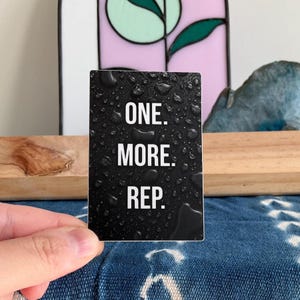 May include: A rectangular sticker with a black background featuring a realistic pattern of water droplets. The white text printed in the center of the sticker reads ONE. MORE. REP. in a clean, sans-serif font, arranged in three stacked lines.