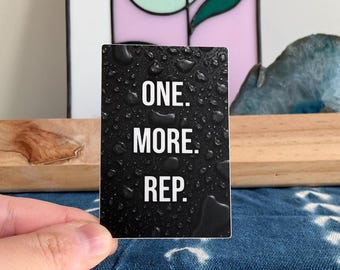 Op de afbeelding: Een rechthoekige sticker met een zwarte achtergrond en een realistisch patroon van waterdruppels. De witte tekst in het midden van de sticker luidt ONE. MORE. REP. in een strak, schreefloos lettertype, gerangschikt in drie regels.