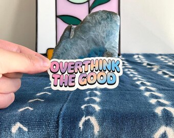 Op de afbeelding: Een gestanste sticker met de tekst OVERTHINK THE GOOD in een bubbelachtig, afgerond lettertype. De letters hebben een kleurverloop van zachtroze naar oranje en lichtblauw, omlijnd met een dikke zwarte rand op een witte achtergrond.