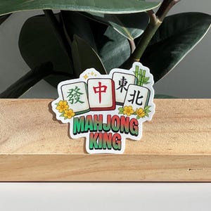 May include: A sticker featuring a Mahjong King design. The sticker showcases four Mahjong tiles with Chinese characters and the words "Mahjong King" in green. Yellow flowers and bamboo stalks decorate the edges of the tiles.