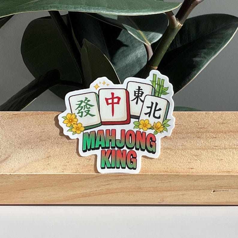May include: A sticker featuring a Mahjong King design. The sticker showcases four Mahjong tiles with Chinese characters and the words "Mahjong King" in green. Yellow flowers and bamboo stalks decorate the edges of the tiles.