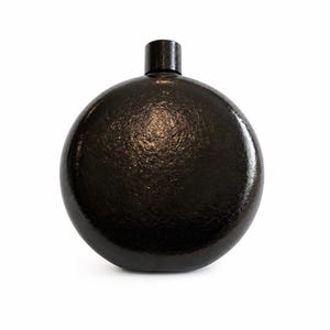 May include: A circular, black flask with a textured, hammered surface finish. The container features a cylindrical neck at the top center, capped with a matching black, smooth, cylindrical stopper. The overall design is symmetrical and rounded.