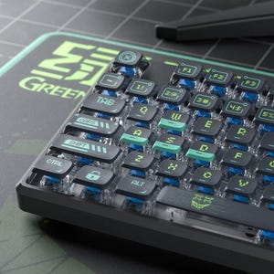 May include: A mechanical keyboard with transparent keycaps and blue and green detailing. The keyboard has a black frame and sits on a green and black mousepad. The keys are labelled with white and green text. The word "GREEN" is visible on the mousepad.