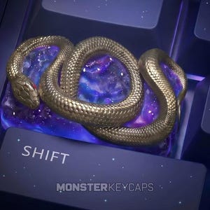 May include: A custom mechanical keyboard keycap featuring a coiled metallic gold snake resting on a translucent purple and blue galaxy-patterned base. The keycap is positioned above a dark blue shift key labeled SHIFT and MONSTERKEYCAPS.