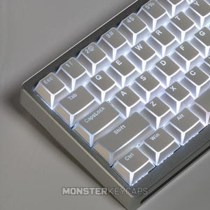 Könnte beinhalten: Eine mechanische Tastatur mit einem silbernen Metallrahmen und weißen Tastenkappen mit schwarzer Beschriftung. Die Tasten sind mit hellem weißem Licht hinterleuchtet, das die Lücken zwischen ihnen beleuchtet. Der Text MONSTERKEYCAPS steht unten.
