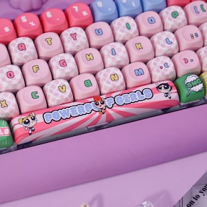 May include: A close-up of a custom keyboard with pink, white, and blue keycaps. The keycaps have letters, numbers, and symbols. The space bar features the text "POWERPUFF GIRLS" with cartoon characters and a pink and white striped design.