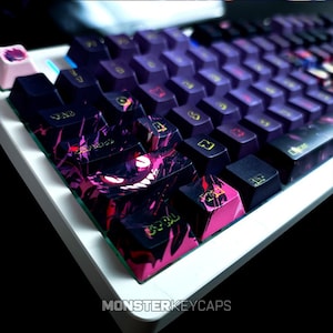 May include: A white mechanical keyboard featuring custom keycaps with a dark purple and black abstract pattern. The caps display a graphic of a grinning creature with glowing eyes on the control key and yellow lettering for the key labels.
