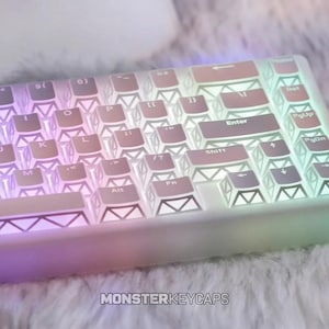 Puede incluir: Un teclado mecánico con teclas translúcidas que presentan un patrón geométrico blanco en los laterales. Las teclas están iluminadas por una suave retroiluminación degradada multicolor, y el texto MONSTERKEYCAPS aparece en la parte inferior.