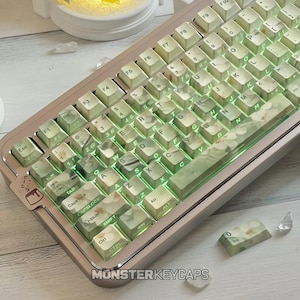 Può includere: Una tastiera meccanica con tasti a motivo floreale verde chiaro e bianco con retroilluminazione. La tastiera ha una cornice metallica e include un tasto abbinato separato. Il testo MONSTERKEYCAPS è visibile nella parte inferiore della cornice.