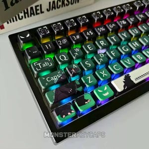 Puede incluir: Un teclado mecánico con teclas personalizadas que presentan gráficos de monstruos, incluyendo marcas de garras, siluetas y ojos verdes brillantes. Las teclas están retroiluminadas con una vibrante iluminación RGB y la barra espaciadora.