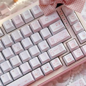 May include: A white and pink mechanical keyboard with custom keycaps. The keycaps feature pink illustrations of characters and symbols. The keyboard has a pink frame and a pink bow in the background. The keyboard has the text "SHIFT" and "CTRL" on the keys.