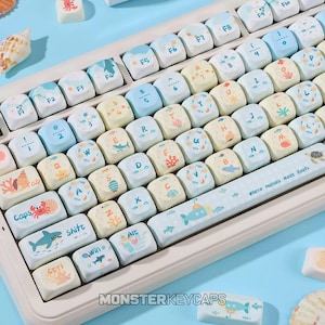 May include: A mechanical keyboard with a light-colored frame and custom keycaps featuring a marine theme. The keys display letters, numbers, and symbols alongside illustrations of sea creatures, coral, and shells. The spacebar reads Where Humans Meet Reefs.