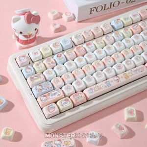 Puede incluir: Un teclado mecánico blanco con teclas de colores pastel que presentan ilustraciones de personajes, corazones y estrellas. La barra espaciadora muestra el texto HELLO KITTY. Una pequeña figura de Hello Kitty se encuentra junto al teclado.