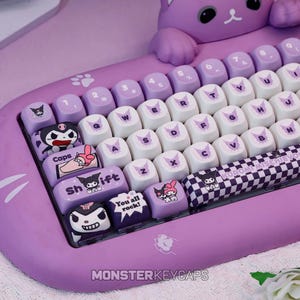 May include: A purple mechanical keyboard featuring a cat-shaped frame and custom keycaps with Kuromi and My Melody character graphics. The keys include text such as Caps, Shift, and You all rock, alongside a checkered spacebar with the text KUROMI MISCHIEVOUS.