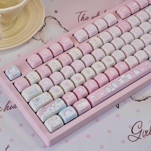 May include: A pink mechanical keyboard with white and pink keycaps. The keycaps feature various illustrations of cute characters. The keyboard has a light pink frame and a matching space bar with a repeating pattern of small characters.