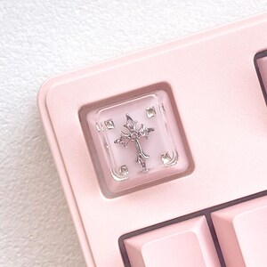 May include: A close-up of a pink keyboard with a custom keycap. The keycap is clear with a silver cross design in the center. The keyboard has a light pink colour scheme.