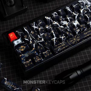 Puede incluir: Un teclado mecánico con teclas personalizadas con patrón de mármol en blanco y negro, letras doradas y símbolos de inspiración nórdica. Incluye una tecla roja con un icono de martillo dorado y una barra espaciadora con arte lineal dorado.