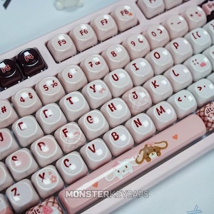 Puede incluir: Un teclado mecánico con teclas redondeadas en color rosa claro y blanco con letras de color rojo oscuro. La barra espaciadora muestra un gráfico de personaje de dibujos animados con un corazón. El texto MONSTERKEYCAPS es visible en la base.