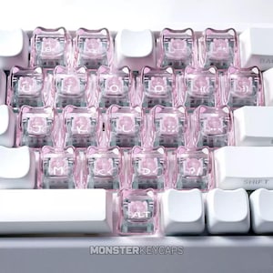 May include: A collection of translucent pink cat-shaped keycaps installed on a mechanical keyboard. Each keycap features a small pink cat face inside the clear casing and white printed letters. The keyboard includes solid white keys and the text MONSTERKEYCAPS.